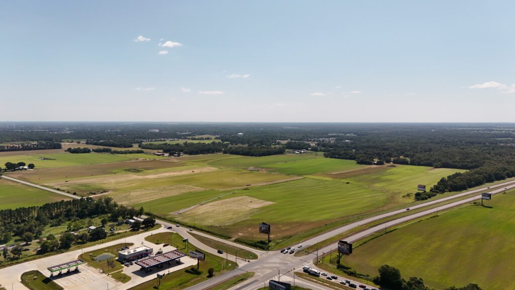 Aerial view of a rural landscape with green fields, a few scattered buildings, and intersecting roads bordered by trees under a clear blue sky. Billboards and vehicles are visible near the road junction in the foreground.