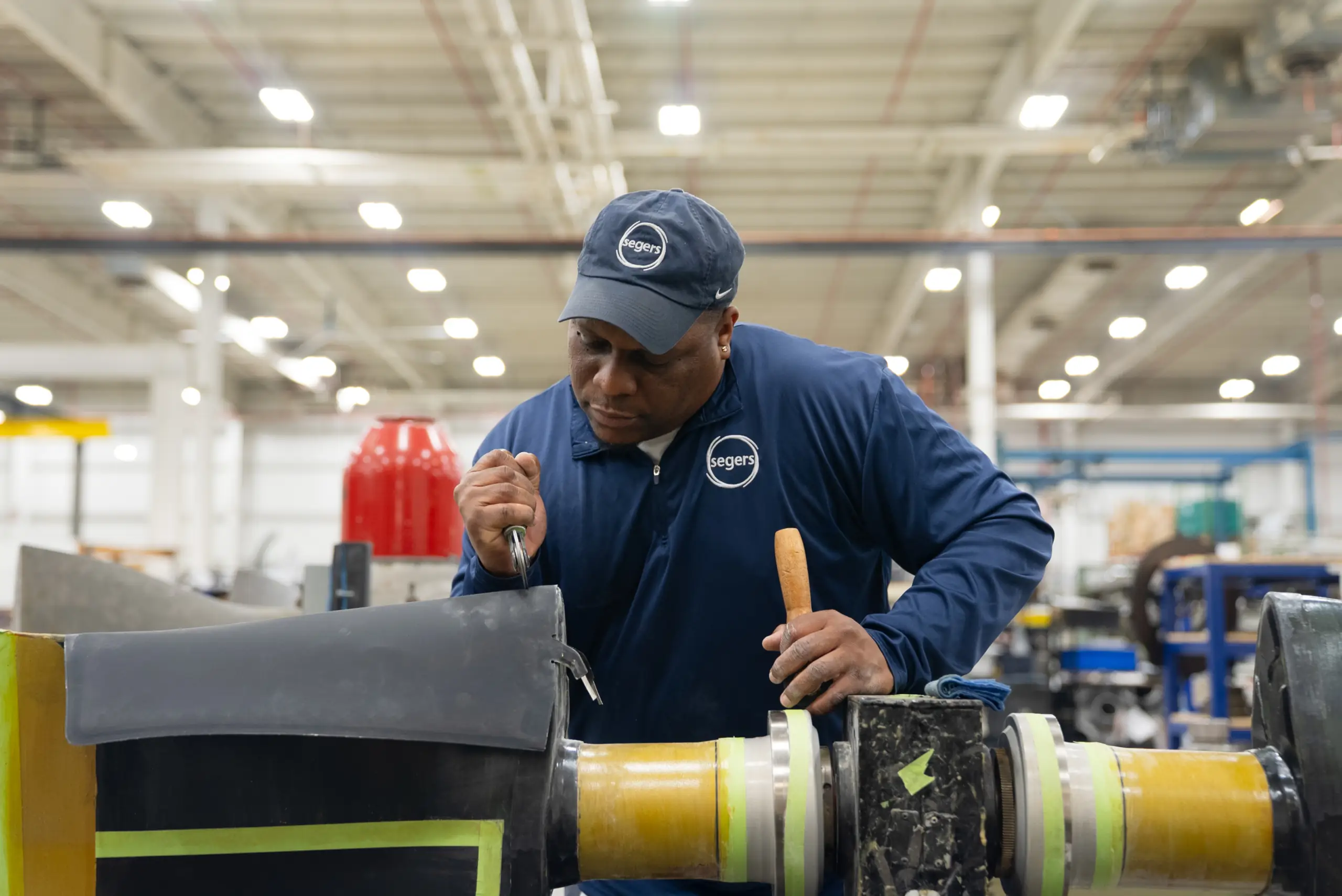 A factory worker in a blue uniform and cap uses a tool to inspect or repair industrial equipment inside a large, well-lit manufacturing facility, similar to the hands-on experience students gain through the Baldwin Prep dual enrollment nursing program at Coastal Alabama.