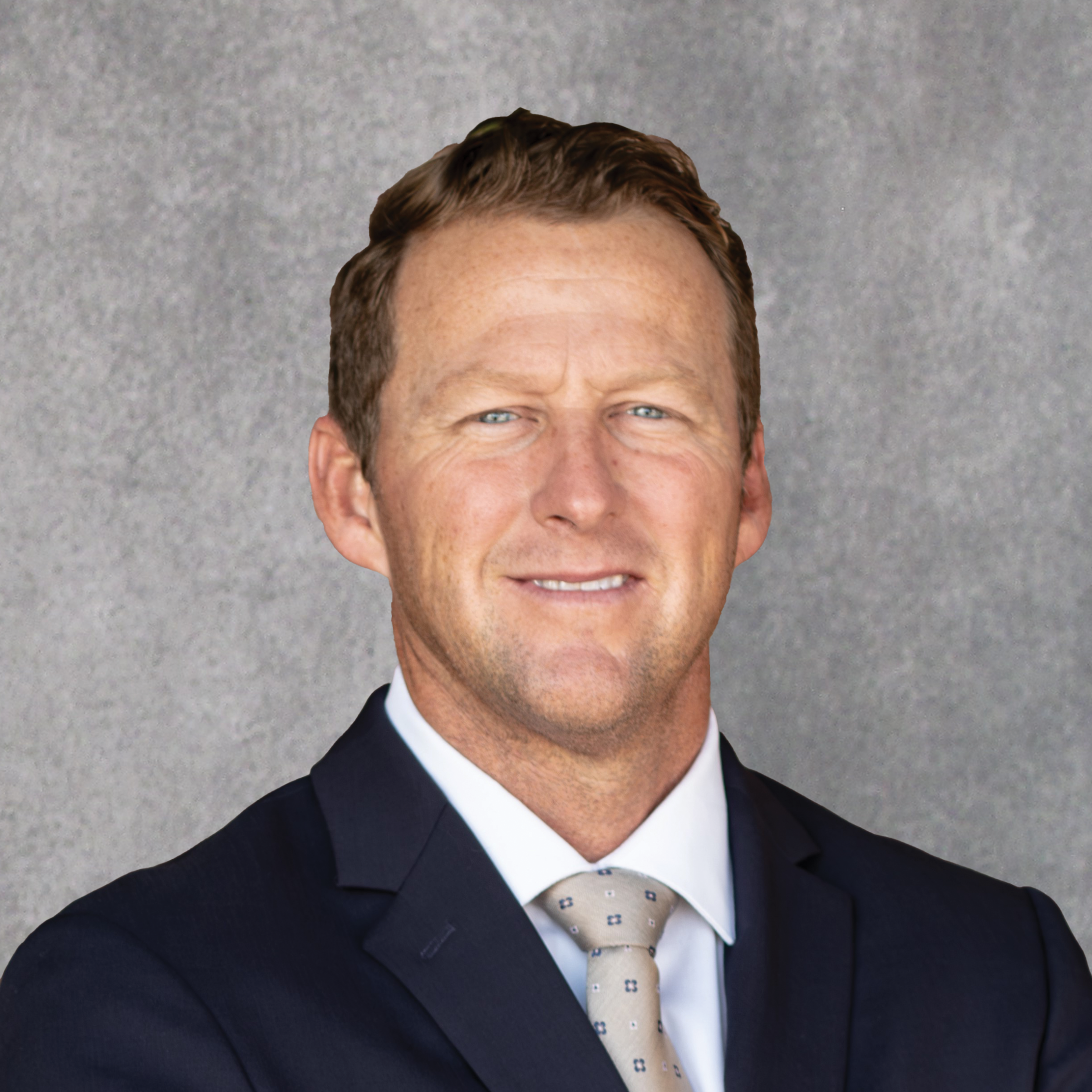 A man with short light brown hair, wearing a dark suit, white shirt, and patterned tie, stands in front of a plain gray background, smiling slightly with arms relaxed at his sides, representing the Baldwin Community & Economic Development Foundation.