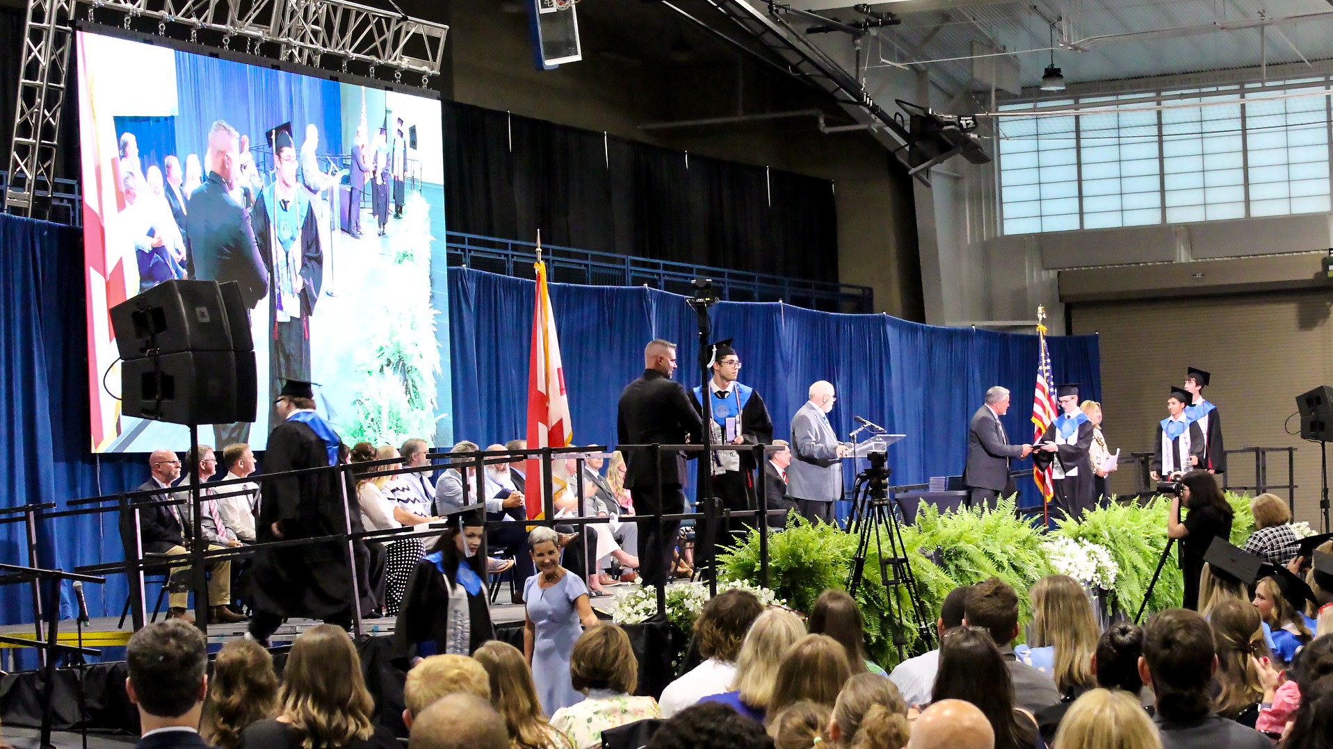 A graduation ceremony takes place indoors with students in caps and gowns on stage. As Buffalo Rock Company expanding to Loxley is announced, graduates receive diplomas from officials. A large screen displays the event, with flags visible in the background.