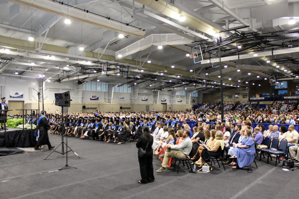 A large crowd gathers in a gymnasium for a graduation ceremony. As Buffalo Rock Company expands to Loxley, excitement fills the air, with graduates in caps and gowns sitting in rows and families filling the audience seats near the stage and microphone.