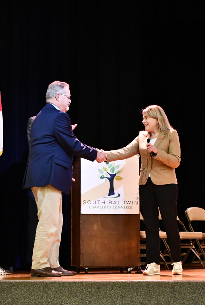 A man and a woman smile and shake hands on stage beside a podium with the South Baldwin Chamber of Commerce logo, celebrating Foley being awarded a $3.3 million SEEDS grant to acquire an industrial park, with empty chairs and a dark background behind them.