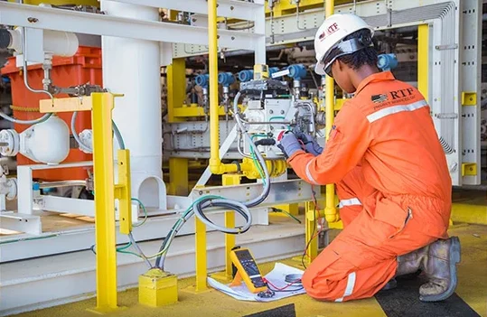 A worker in orange coveralls and a white helmet uses tools and electronic instruments to inspect industrial equipment at a worksite with yellow safety barriers, exemplifying the commitment of an Alabama electrical services company serving energy and industrial industries in Baldwin County, Bay Minette, AL.