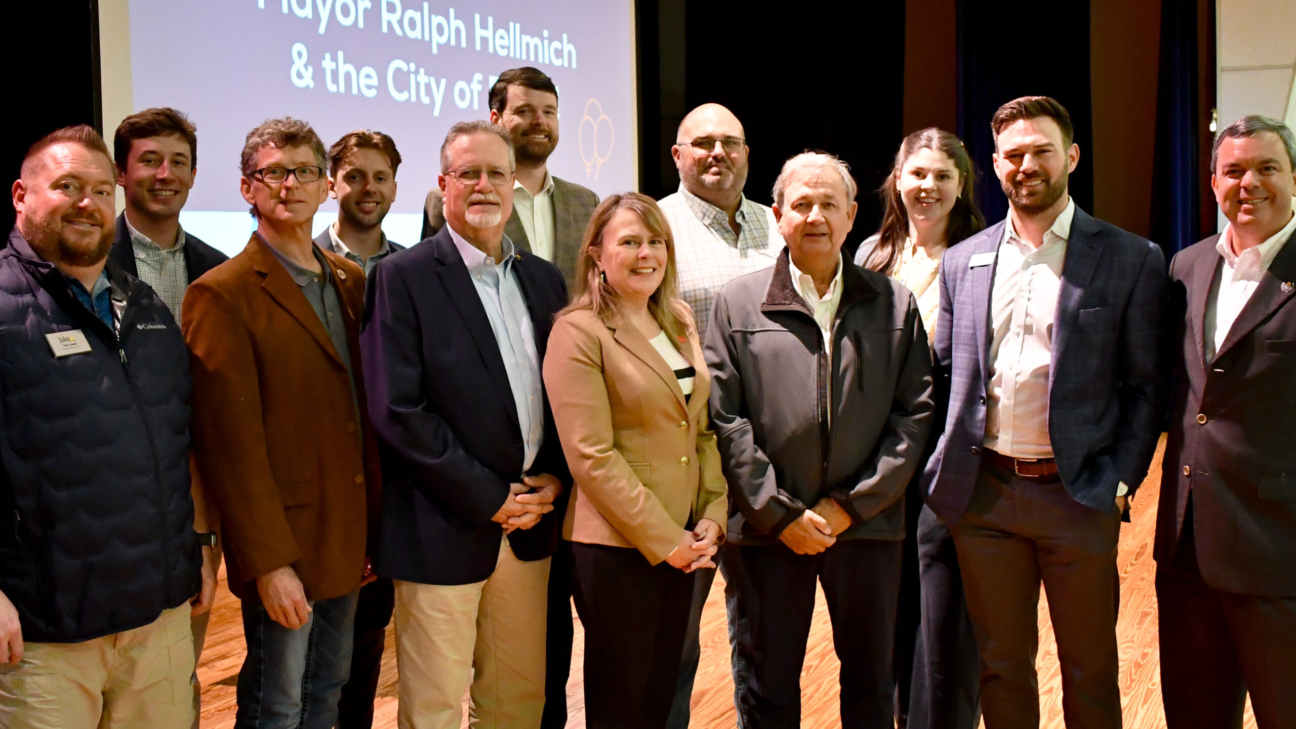 A group of twelve people, dressed in business and casual attire, stand together smiling on a wooden stage. A projection behind them reads Mayor Ralph Hellmich & the City of Foley Awarded $3.3 Million SEEDS Grant to Acquire Industrial Park.