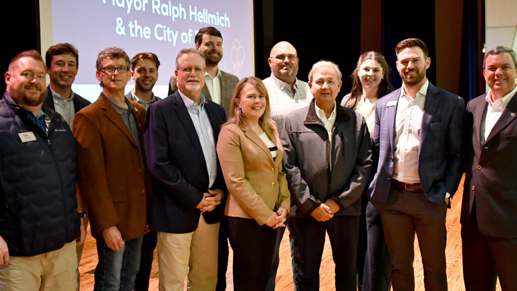 A group of twelve people, dressed in business or business-casual attire, stand smiling together on a stage with a projector screen in the background that reads “Mayor Ralph Hellmich & the City of,” celebrating Foley being awarded a $3.3 Million SEEDS Grant to acquire an industrial park.