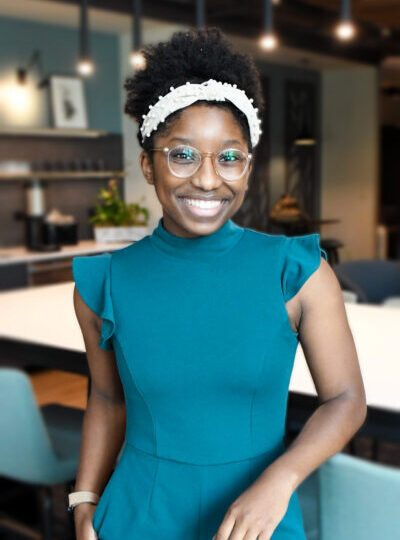 A woman wearing glasses and a teal jumpsuit smiles while standing in a modern office or kitchen space, with chairs and tables in the background.