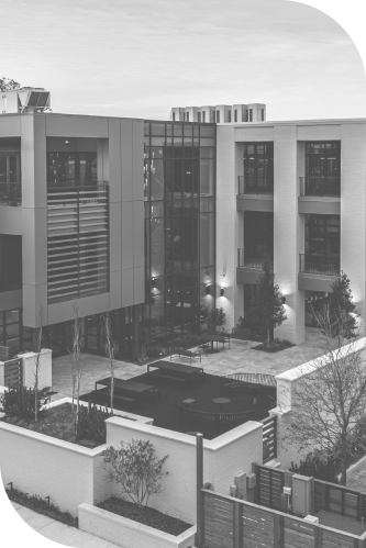A modern three-story building with large glass windows, small balconies, and surrounding landscaping stands prominently. In the foreground, Baldwin Alliance staff can often be seen utilizing a courtyard adorned with benches and planters. The photo is in black and white.