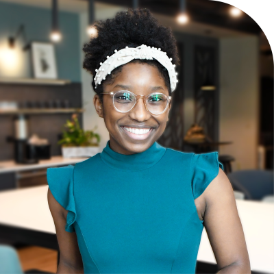 A woman with glasses and a white headband, part of the Baldwin Alliance staff, smiles in a modern office setting, wearing a teal top with ruffled sleeves.