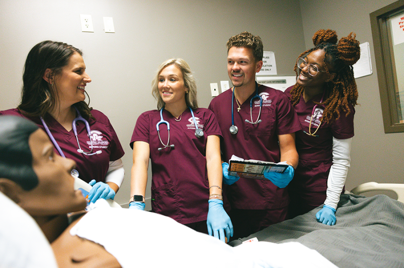 Four nursing students in maroon scrubs stand by a medical mannequin in a hospital bed, smiling as they practice patient care—a scene reminiscent of hands-on training at the 121 acre industrial park site in Foley, Baldwin County, AL.
